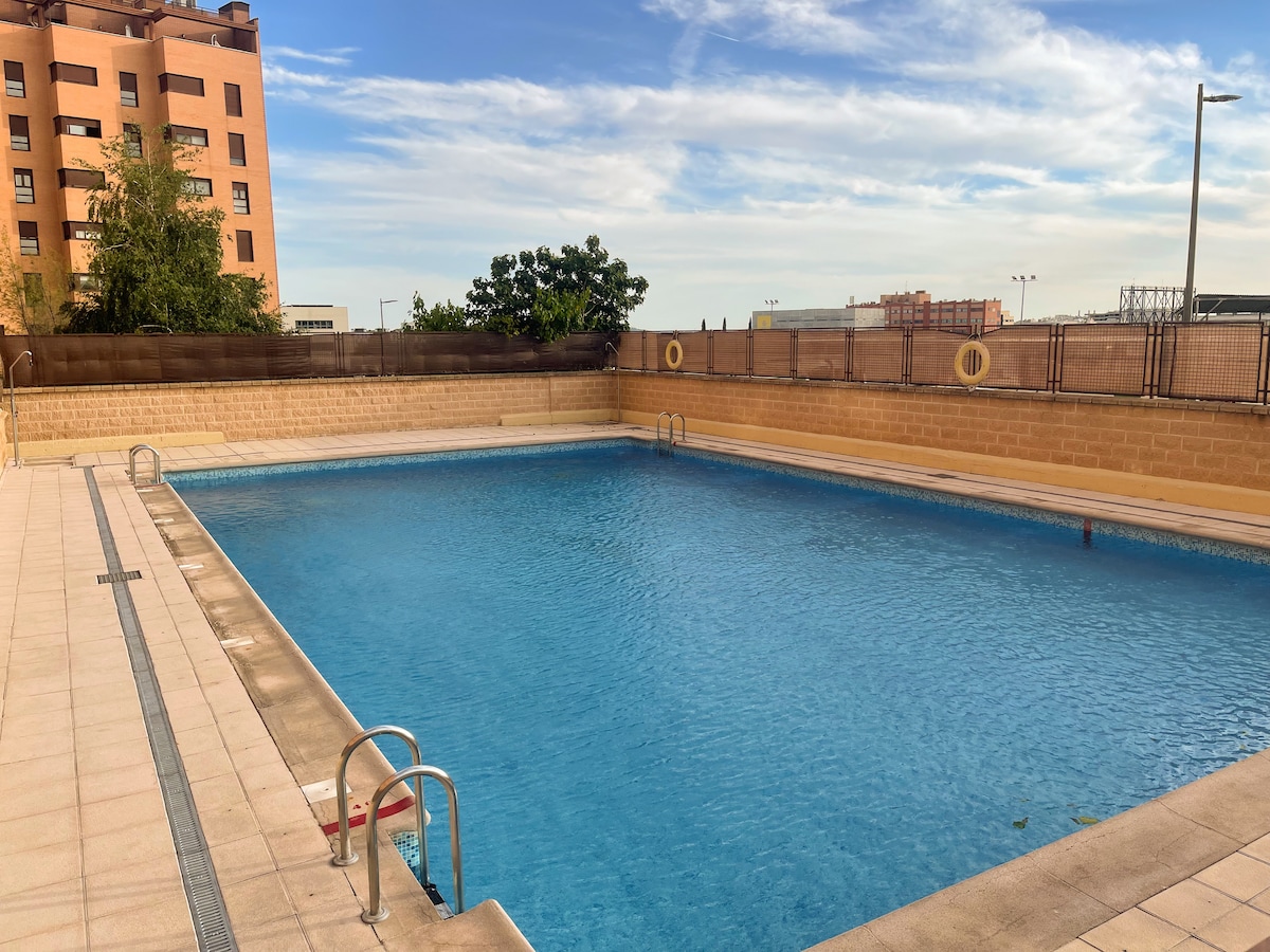 A spacious outdoor swimming pool is presented, featuring clear blue water surrounded by a tiled deck. Sunlight reflects off the surface, enhancing the inviting appearance. Vertical fencing provides a secure boundary, while a few trees and buildings are visible in the background.