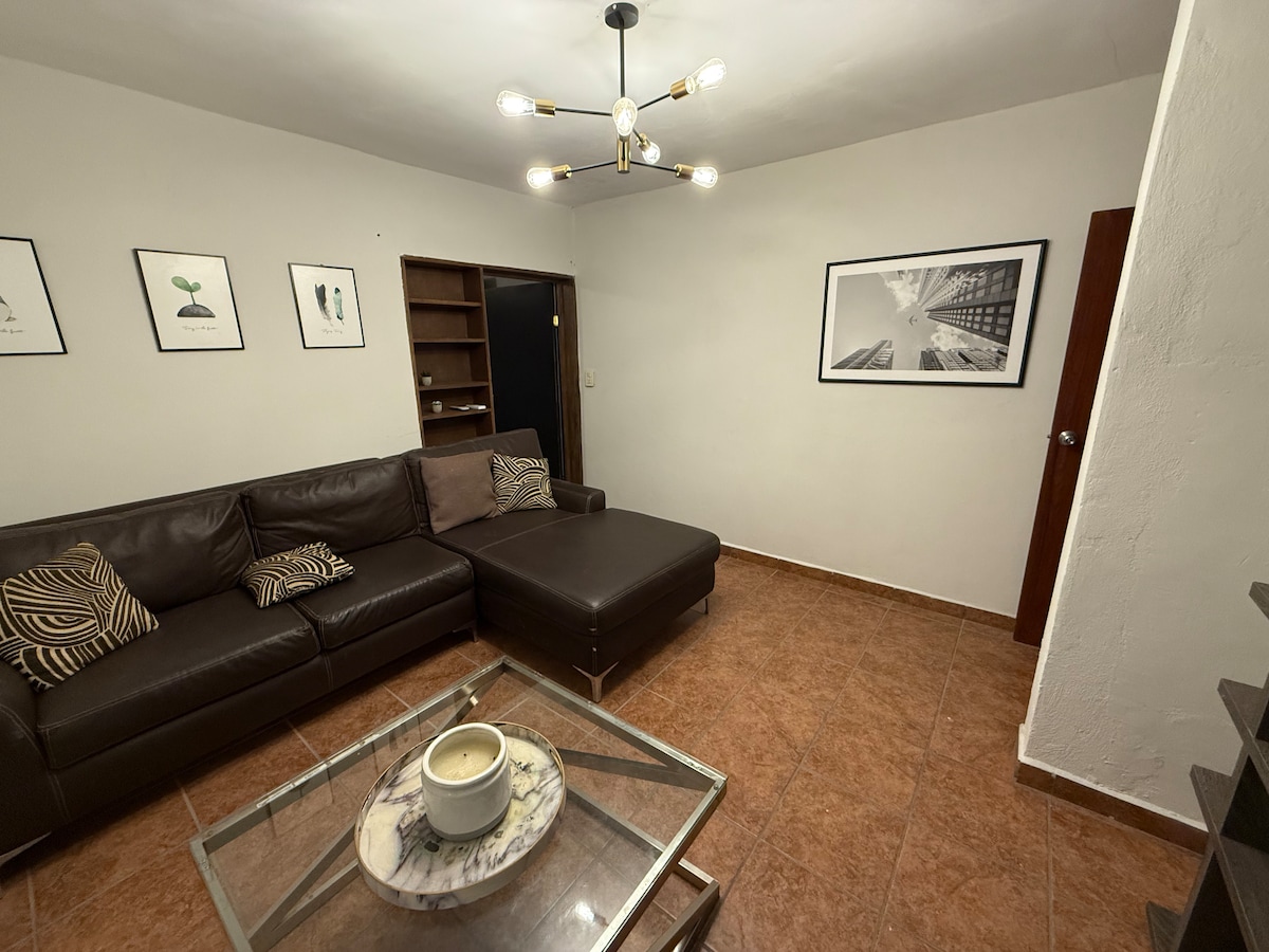 A spacious living area is depicted, featuring a modern sectional sofa and a coffee table with a decorative centerpiece. Art pieces are showcased on the walls, and a bookshelf is visible in the background. The warm lighting enhances the inviting ambiance of the room.