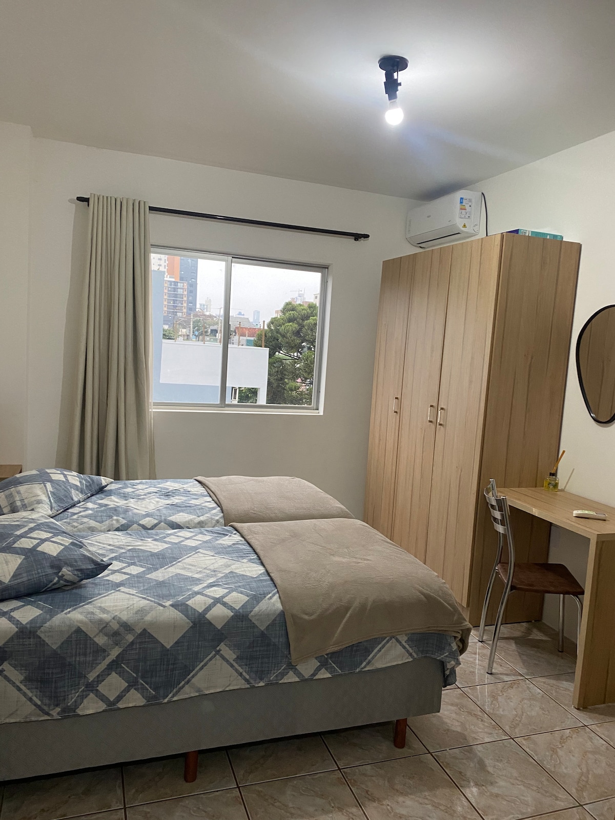 A bedroom is furnished with a spacious bed featuring a patterned blanket. A large window allows natural light to enter, complemented by light-colored curtains. A wooden wardrobe and a round mirror are present, along with a small desk and chair for convenience.