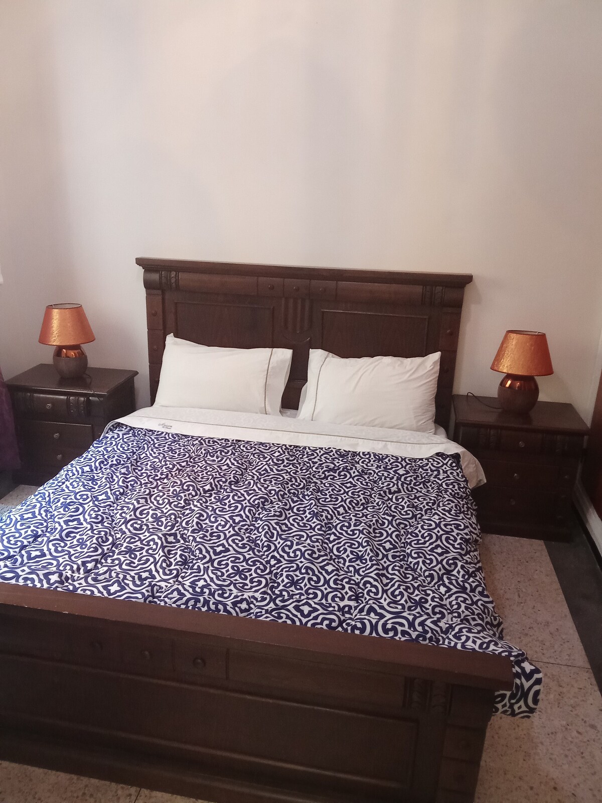 A spacious bed is positioned against a light wall, covered with a patterned blue and white blanket. Two bedside tables with warm-toned lamps sit on either side, providing a cozy touch to the sleeping area.
