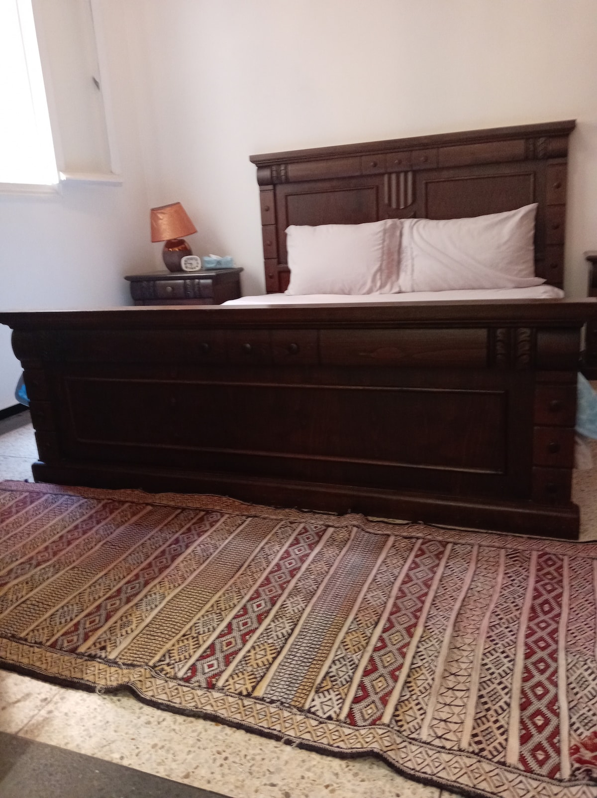 A spacious bedroom features a large wooden bed with a gently carved headboard and neatly arranged light-colored bedding. A bedside table holds a lamp that casts a soft glow. A patterned area rug enhances the cozy feel, placed in front of the bed.