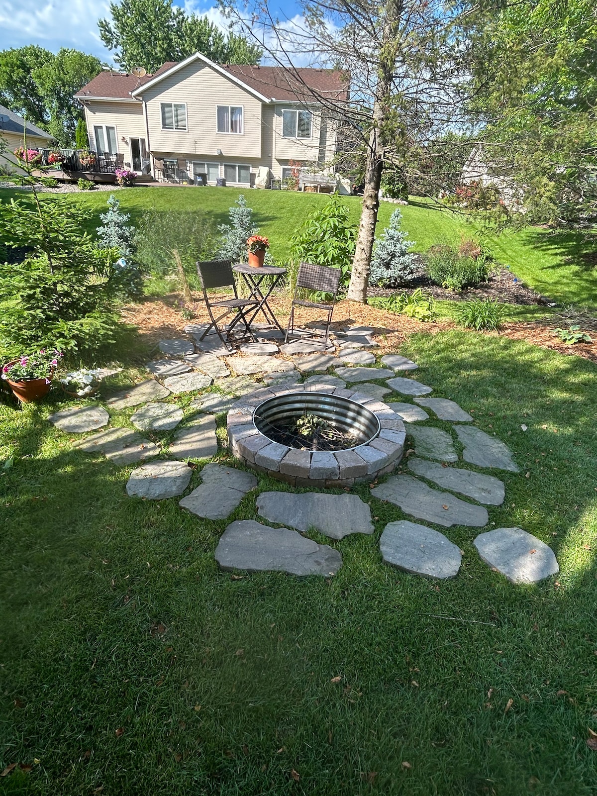 A circular stone patio surrounds a fire pit, with two black chairs positioned nearby. Lush green grass and decorative plants create a serene outdoor space, while a landscaped yard is visible in the background, enhancing a peaceful atmosphere.
