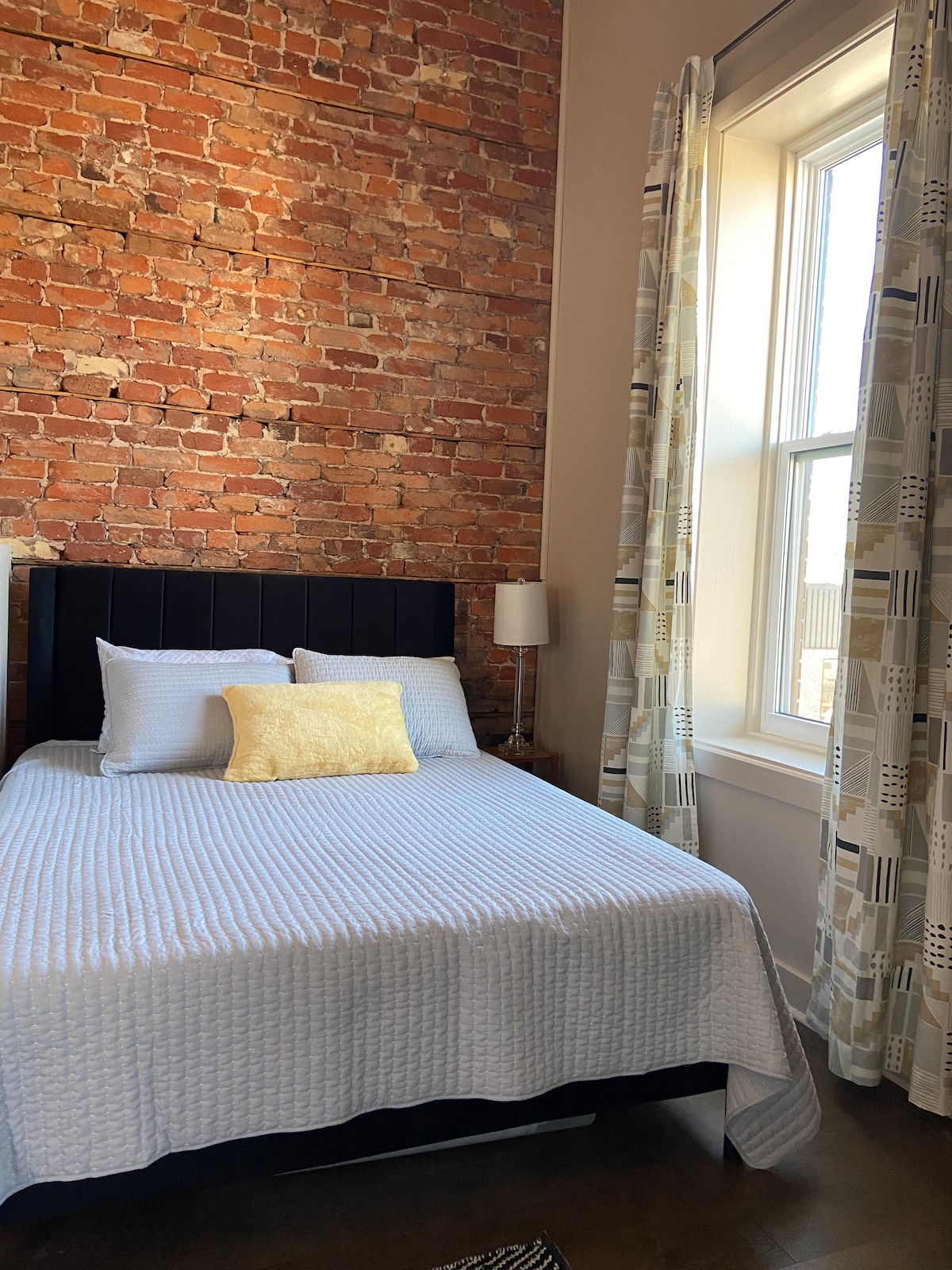 A cozy sleeping area showcases a queen bed with soft bedding and a yellow accent pillow. Exposed brick walls add character, while a window with patterned curtains allows natural light to brighten the space. A bedside lamp complements the minimalist decor.