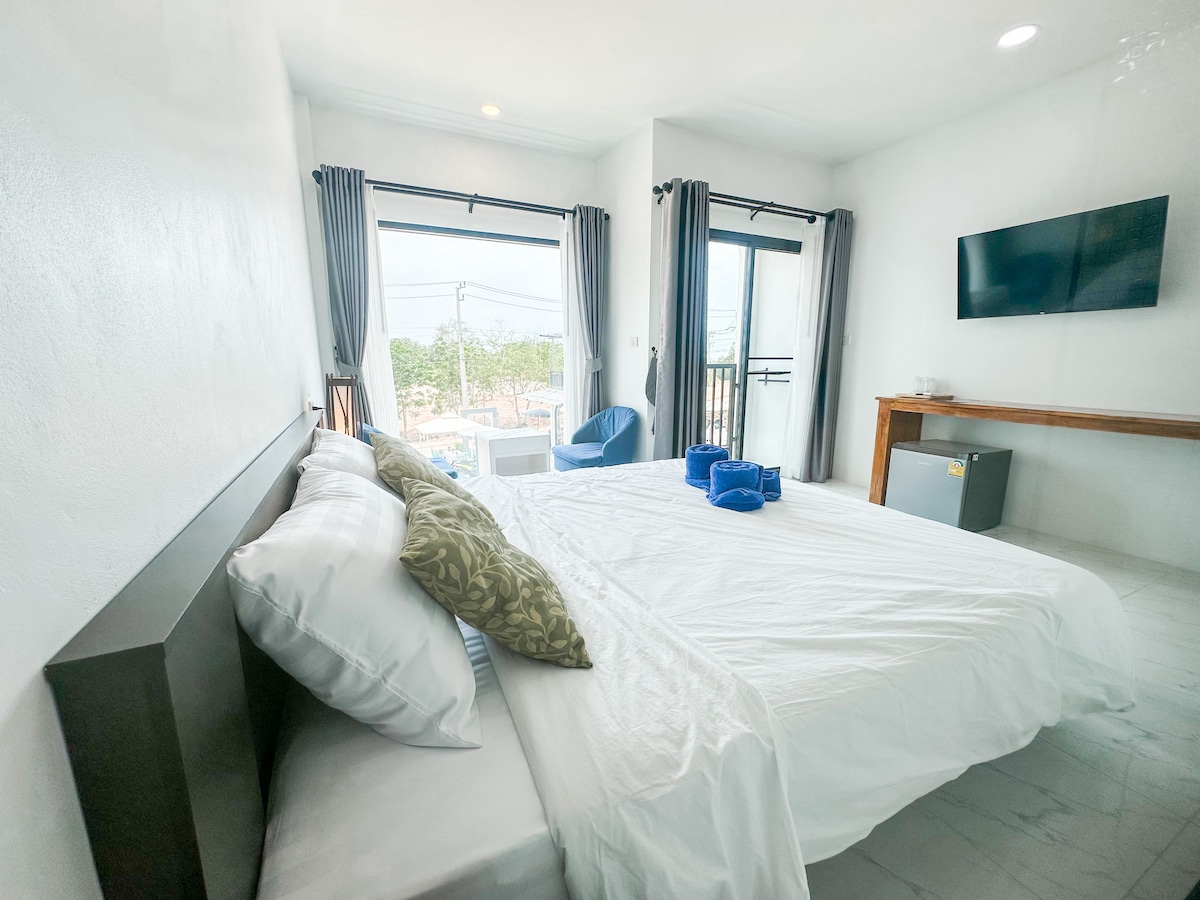 A spacious guest room is furnished with a king bed, featuring a decorative pillow. Large windows allow natural light and views of the surroundings, while a seating area includes a blue chair. A flat-screen TV is mounted on the wall, complementing the minimalist aesthetic.