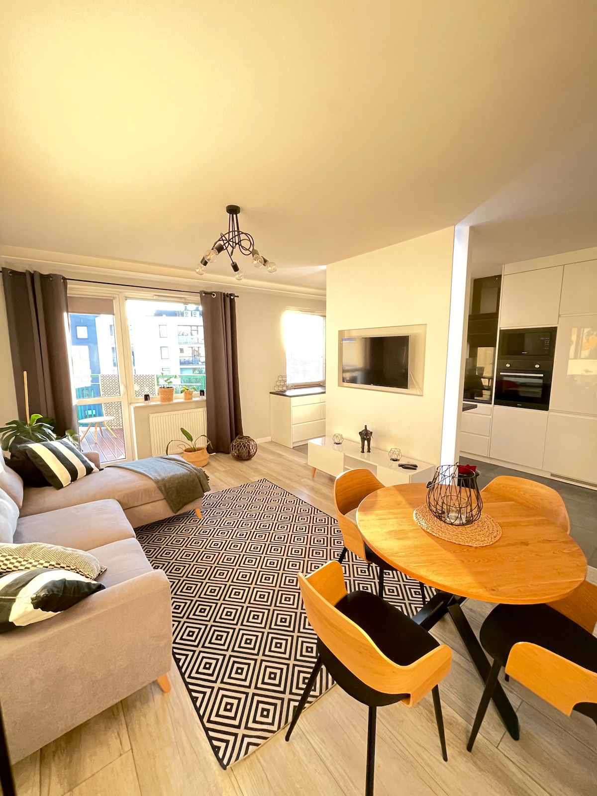 A bright living area features a spacious grey sofa paired with a patterned rug. A round dining table is surrounded by four wooden chairs. Large windows allow natural light to fill the room, while modern decor adds a contemporary touch.