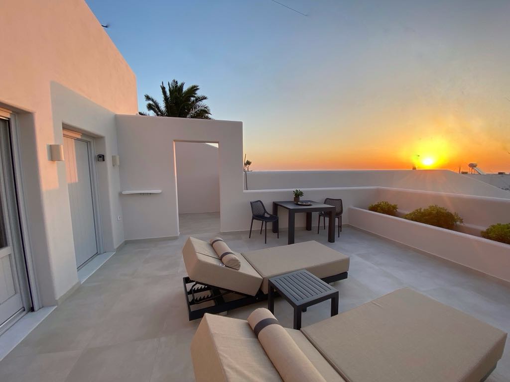 A spacious outdoor terrace is showcased, featuring two lounge chairs and a small table set for four. A sunset casts a warm glow, reflecting against the soft white walls and highlighting the surrounding greenery.