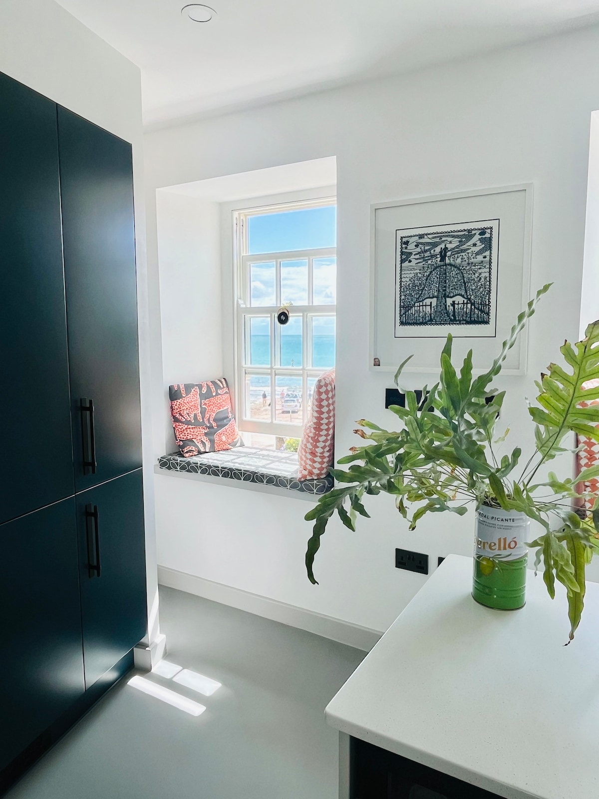 A cozy corner features a window seat adorned with patterned cushions, offering a serene view of the sea. Bright light filters through the window, illuminating the sleek cabinetry and minimalist countertop in the kitchen area, accentuated by a green plant in a decorative vase.