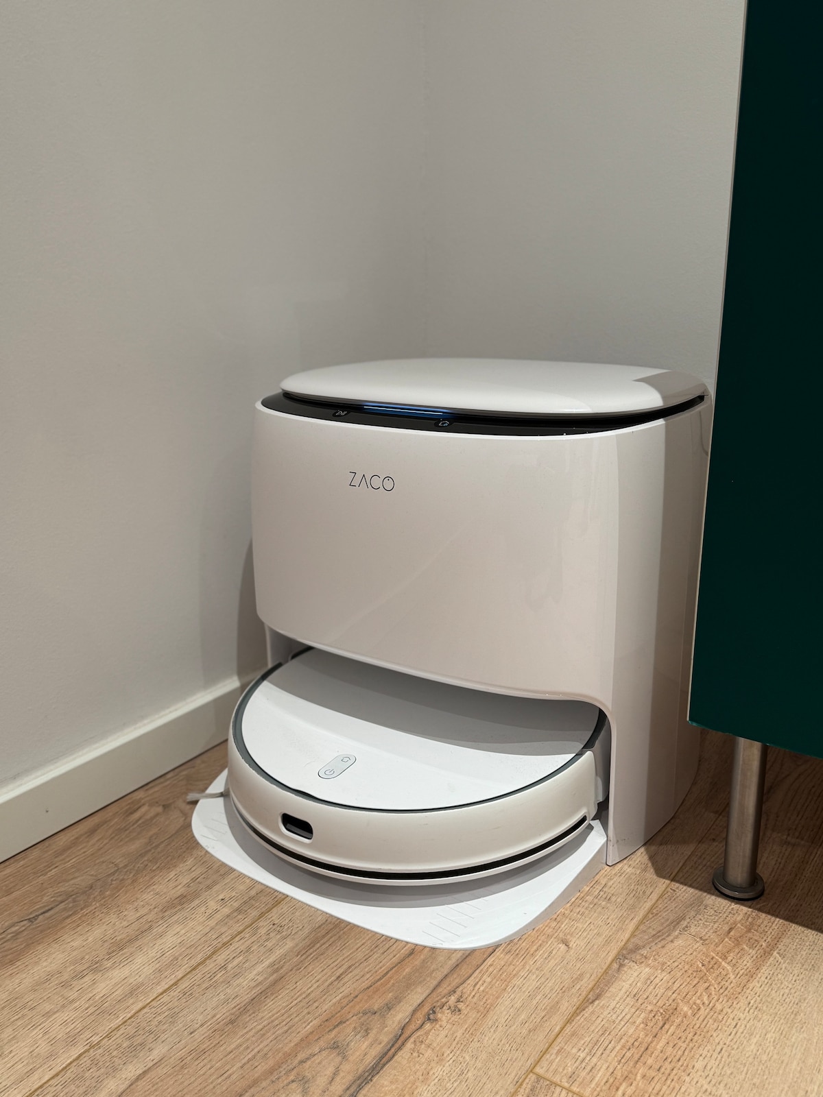 A sleek white robotic vacuum is positioned beside a dark green cabinet on light wooden flooring. The unit has a minimalistic design, featuring a top compartment with a lid and a lower round section designed to move about. Natural light softly illuminates the area.