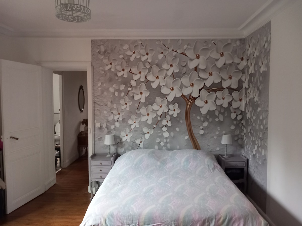 A spacious sleeping area features a large bed with a patterned duvet cover. A mural of white flowers is depicted on the feature wall, complemented by two bedside tables with lamps. A doorway leads to another room, softly illuminated by natural light.