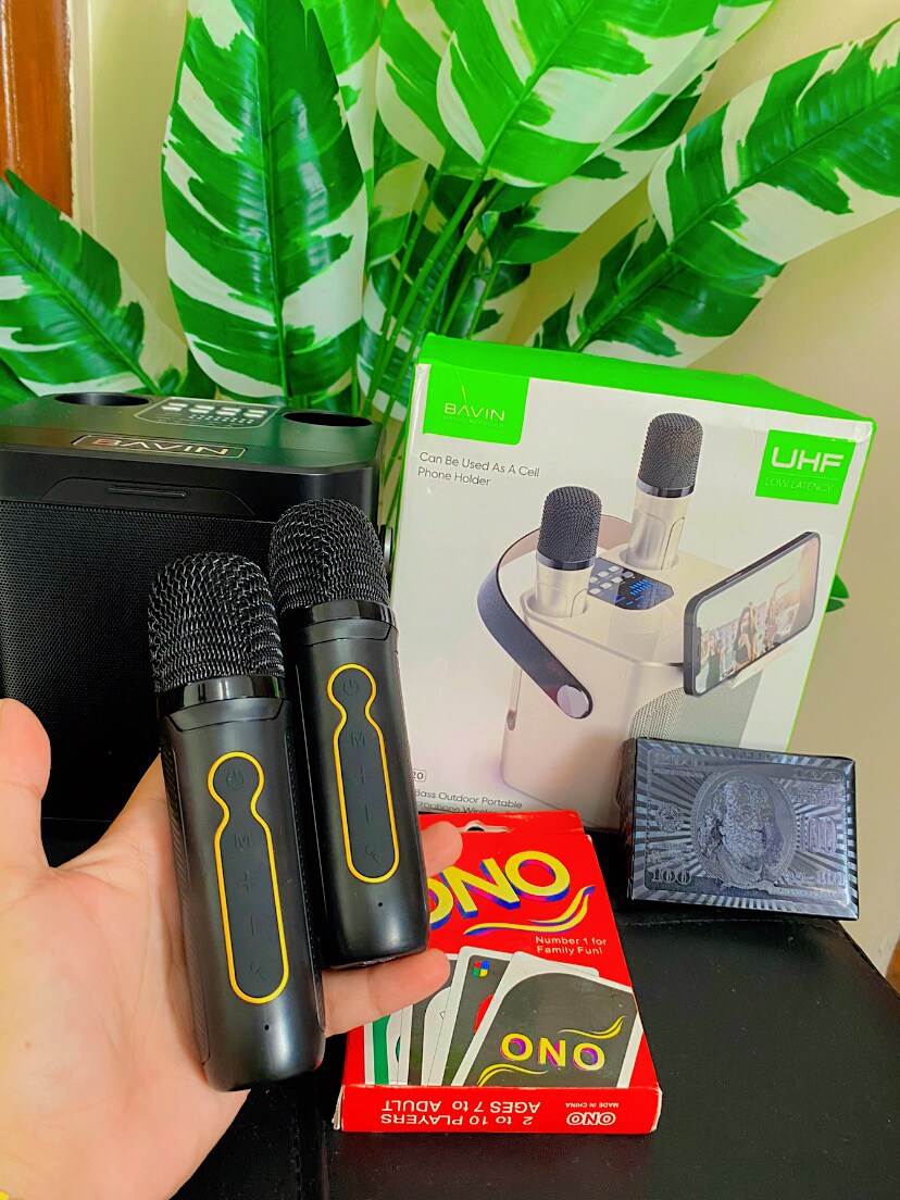 A pair of handheld karaoke microphones rests in a hand, surrounded by a package that includes a microphone holder for mobile devices. A deck of UNO cards and a black decorative item are also visible, creating an inviting setup for social gatherings.