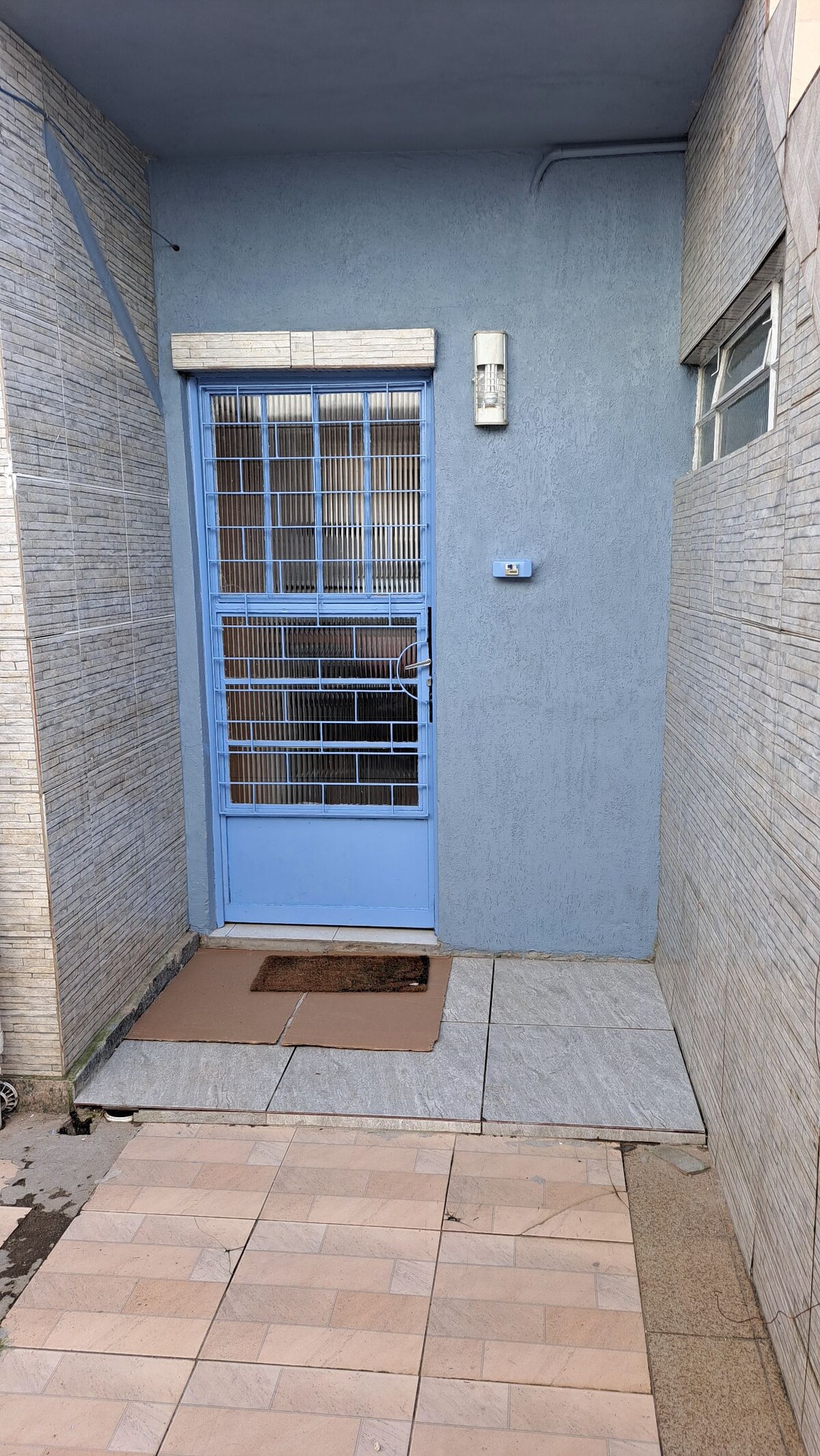 A blue door framed by a light-colored wall stands at the entrance. A small doormat is placed on the tiled floor, leading to the entrance. Natural light illuminates the area, highlighting the textured wall and smooth tiles.