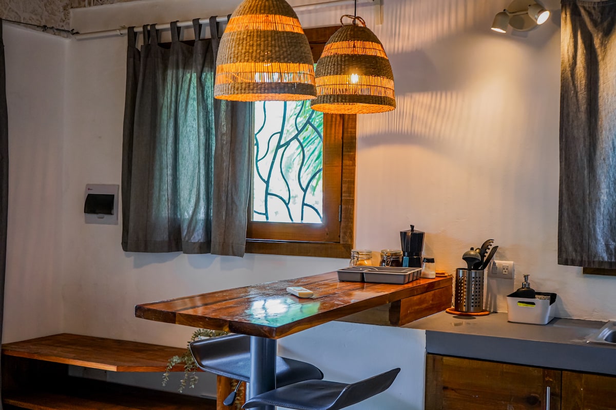 A modern kitchen area is highlighted by a wooden countertop and stylish bar stools. Two woven pendant lights hang above, illuminating the space. A window with stained glass design allows natural light to filter in, complemented by gray curtains on either side.