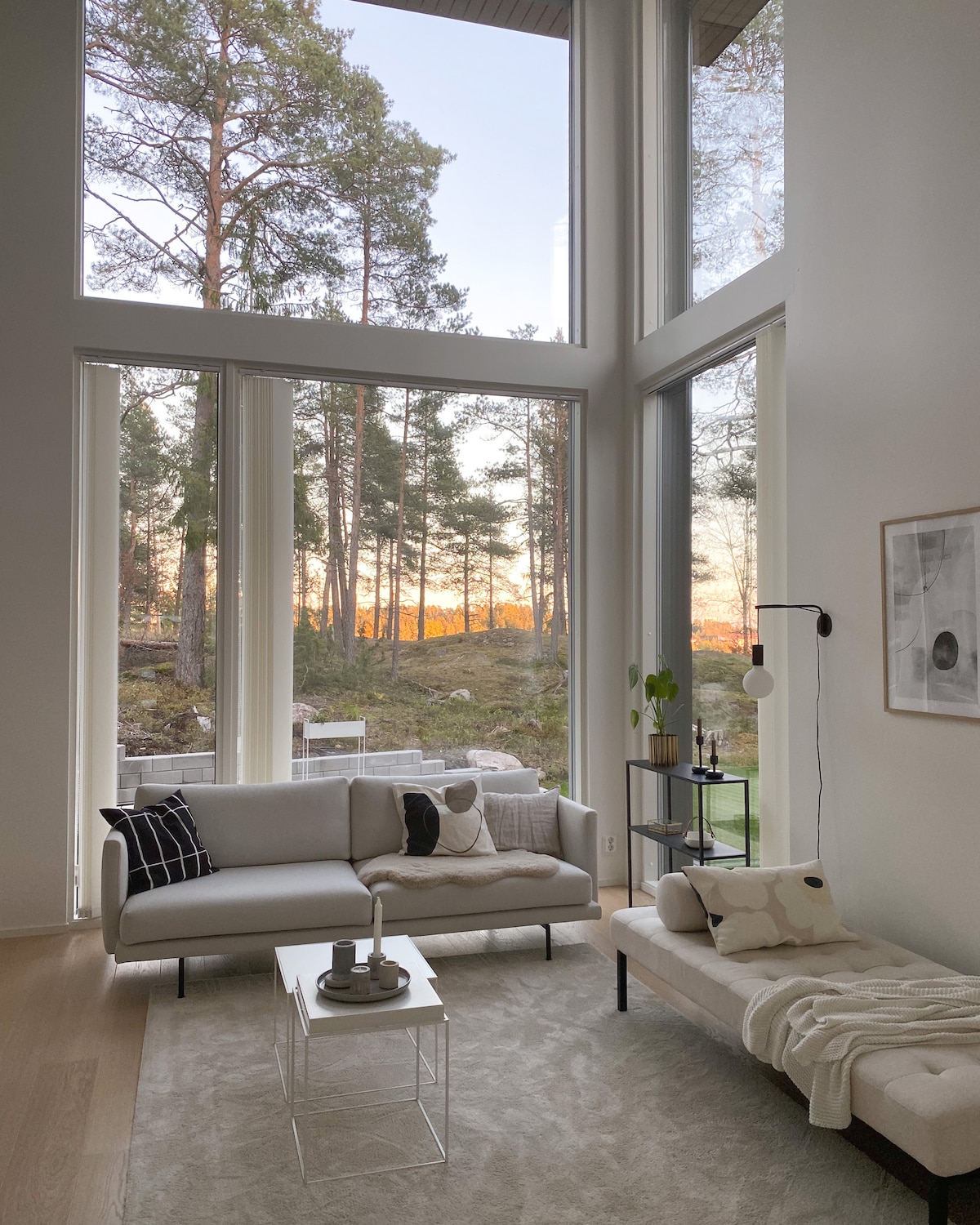 A bright living area features large floor-to-ceiling windows that allow natural light to fill the space. A comfortable sofa and a chaise lounge are positioned near the window, offering views of a forested landscape. A minimalist coffee table and decorative accessories complete the inviting setting.
