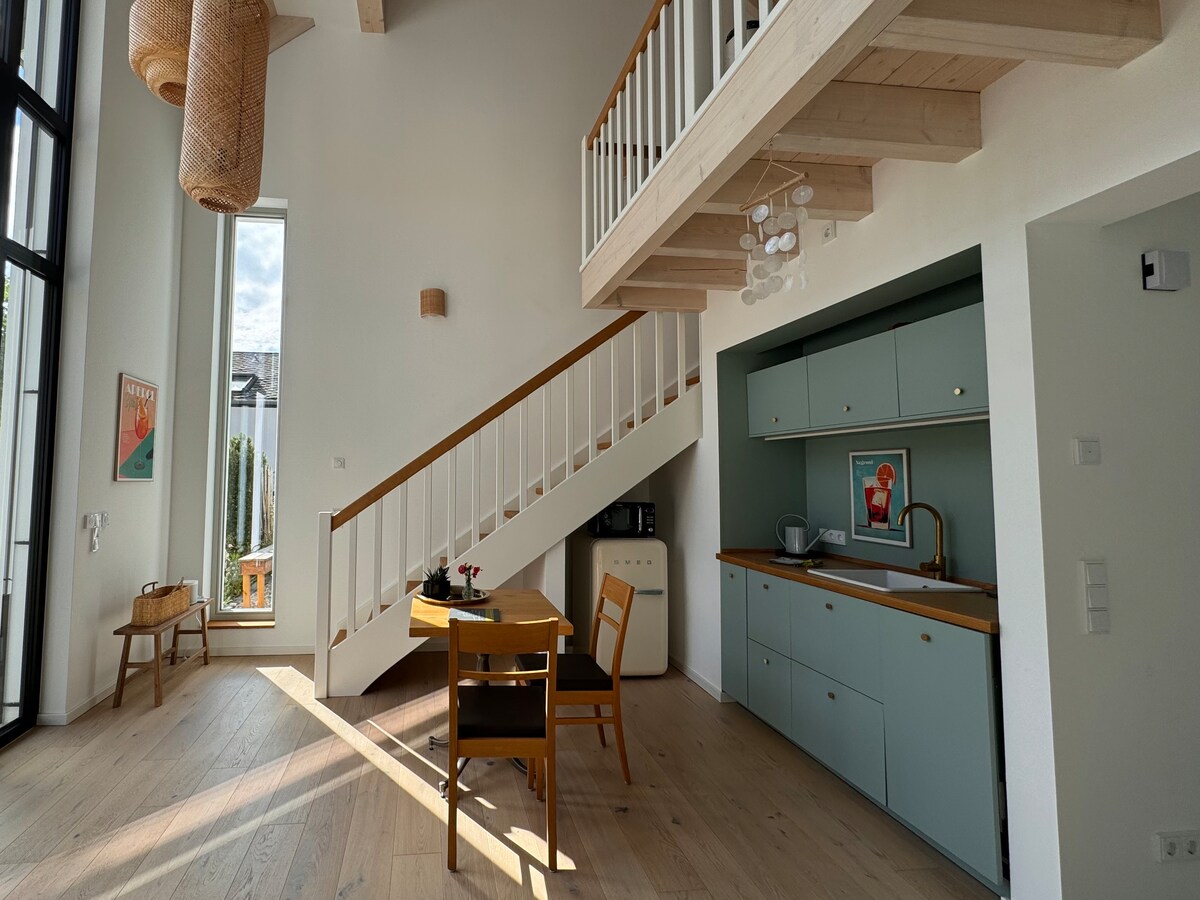 A light-filled open living space features a modern kitchen with pastel green cabinetry and a wooden countertop. A dining table is paired with wooden chairs, and large windows allow natural light to illuminate the room. A staircase leads to a gallery sleeping area above.