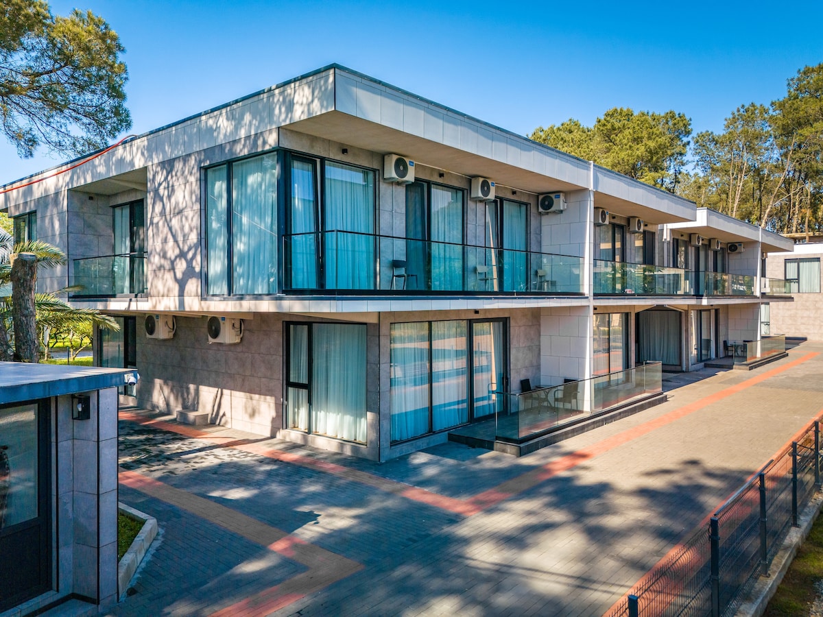 A modern two-story building is viewed from the front, featuring multiple balconies with glass railings. Air conditioning units are installed on the exterior walls. Lush greenery surrounds the property, and a paved path leads to the entrance.