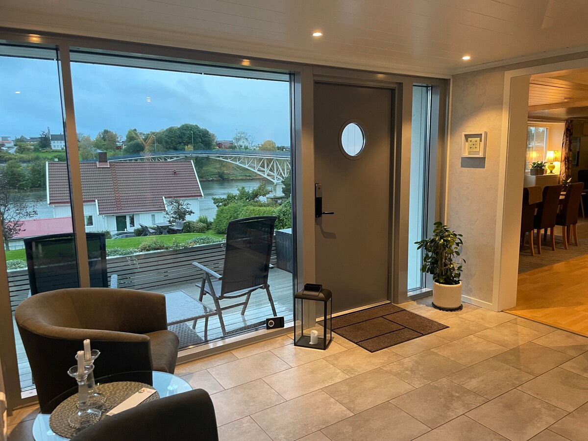 A welcoming entrance features large windows that provide views of the exterior deck and waterway. A modern chair and small table are situated on the tiled floor, with a potted plant adding a touch of greenery. Soft lighting enhances the inviting atmosphere.