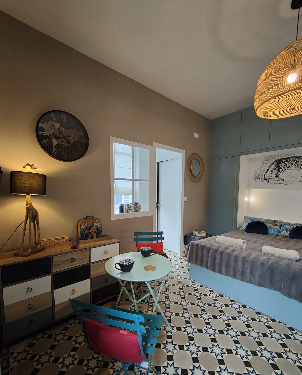The studio features a stylish decor with a cozy sleeping area alongside a small table and chairs. The room is accented by patterned tile flooring and a light fixture with a natural weave. A decorative wall art piece is visible above the bed, enhancing the space's charm.