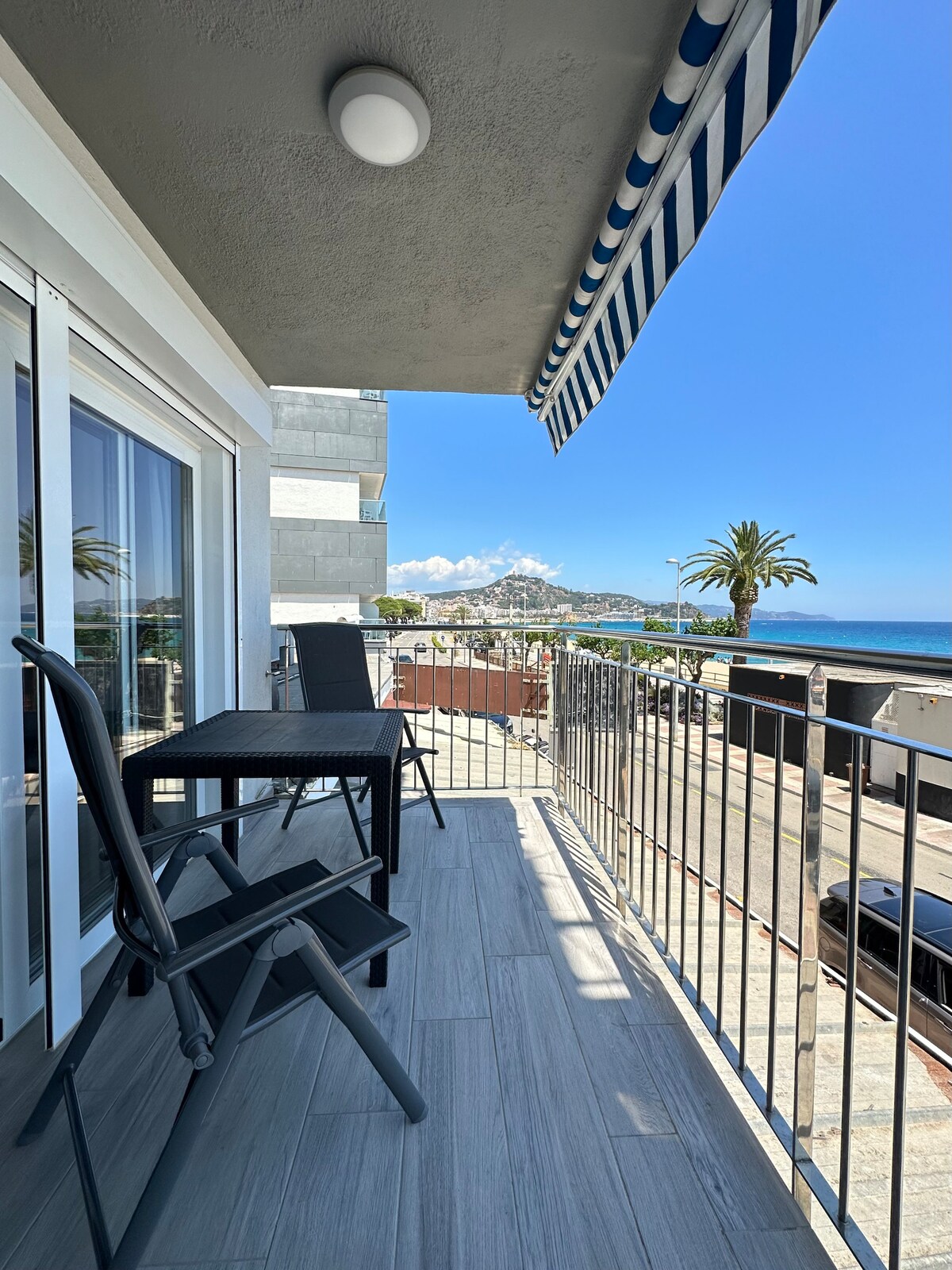 A spacious balcony is presented, featuring two black chairs and a small table. A white awning provides shading, while a view of the ocean and palm trees is visible in the background. Clear skies enhance the coastal setting.