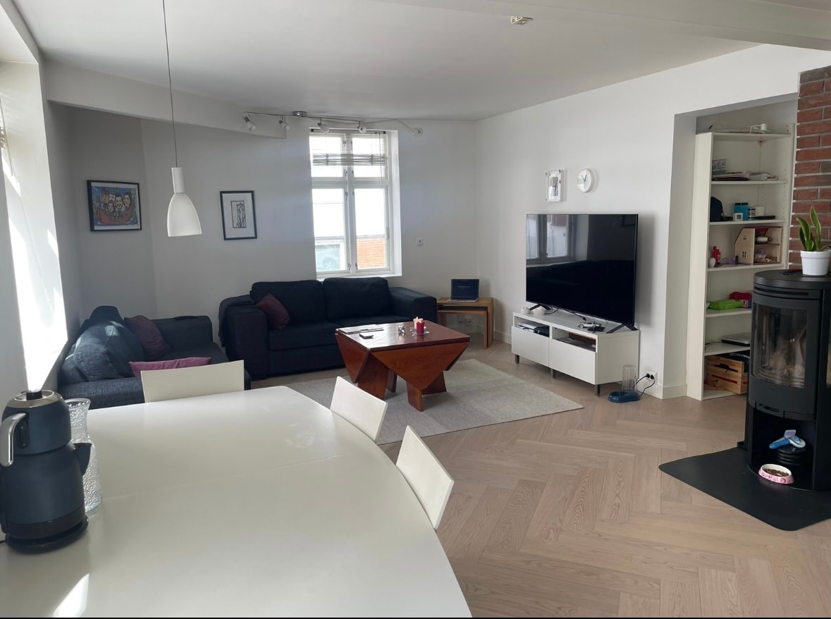 A spacious living area displays a dark sectional sofa facing a modern television on a white media console. A wooden coffee table sits centrally, complemented by a white dining table set for four. Natural light enters through large windows, enhancing the light wood flooring.