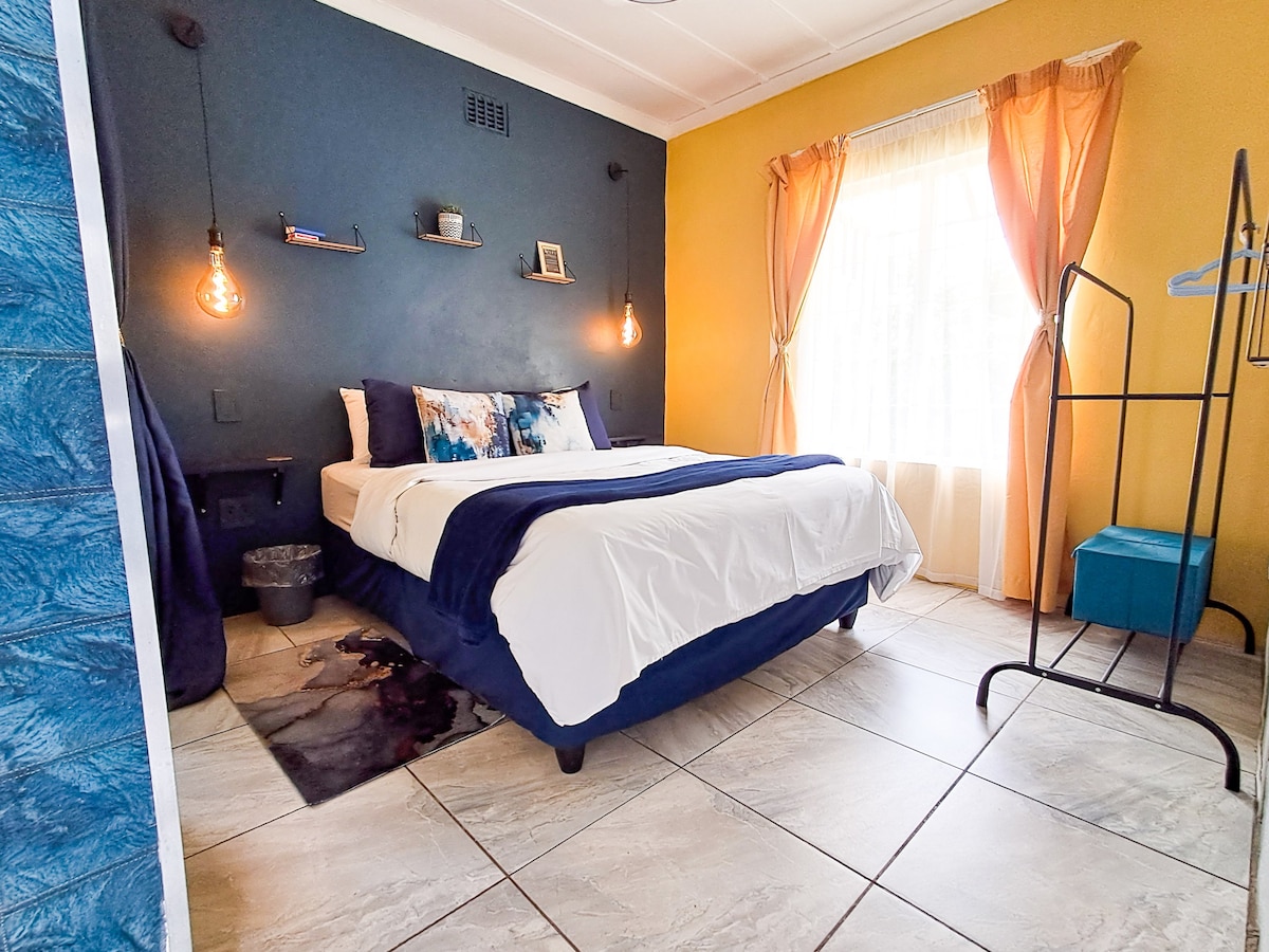 A cozy bedroom features a queen-size bed dressed in white linens and accented with blue covers. The room has a blend of yellow and blue walls, along with hanging pendant lights. A window allows natural light, and a wardrobe stands in the corner.