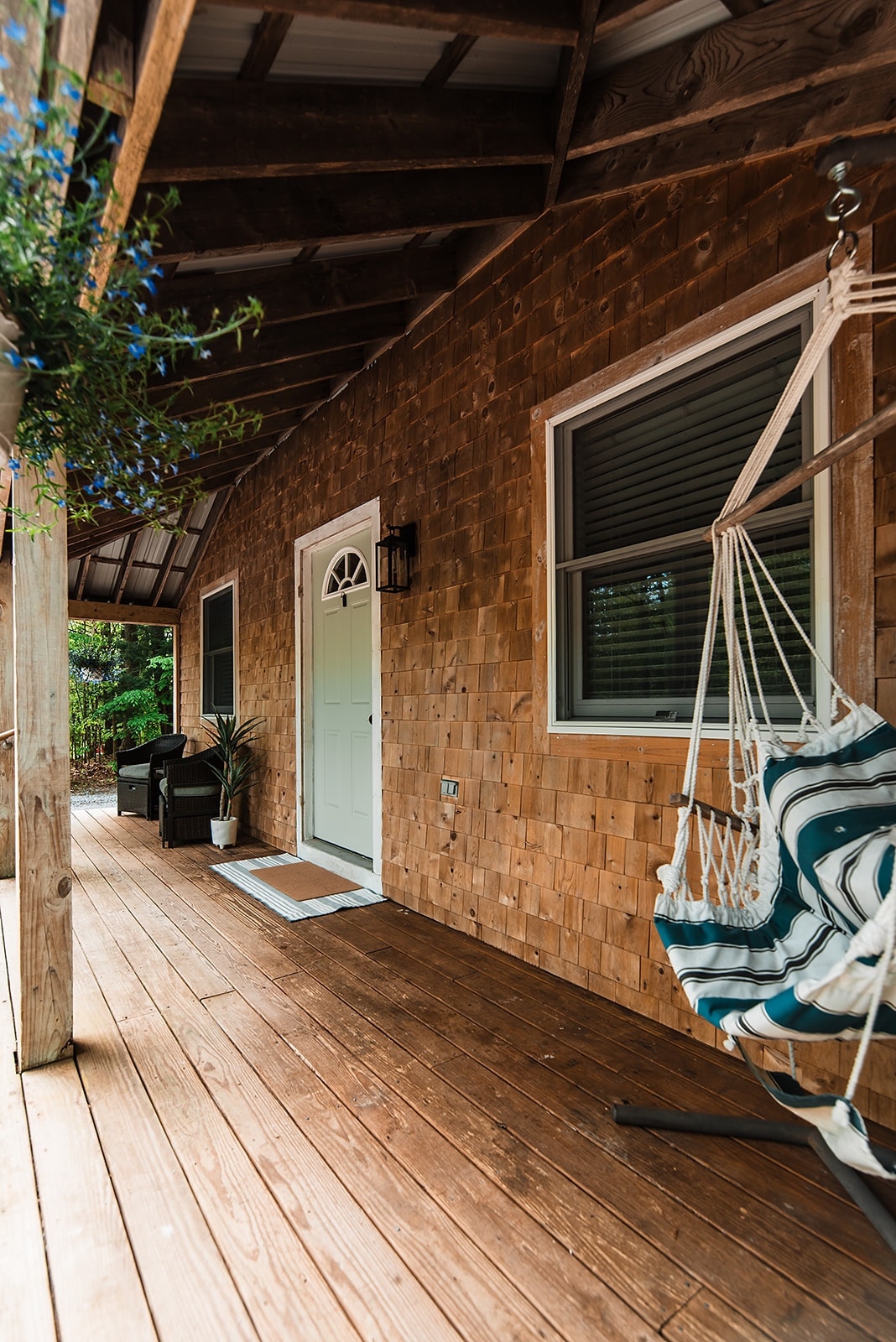 A covered porch is presented with wooden decking and walls made of natural shingles. A white door serves as the main entrance, complemented by a chair and a hanging swing chair with a striped cushion. Green plants and a blue floral arrangement add subtle color.