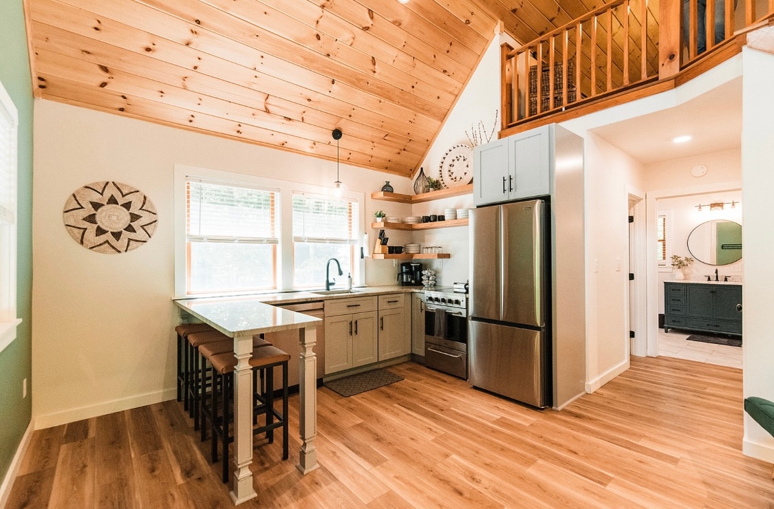 The kitchen area features a wooden ceiling that adds warmth to the space. A modern island with seating for four is complemented by stainless steel appliances, including a refrigerator. Shelves showcase decorative items, while natural light filters through the windows.