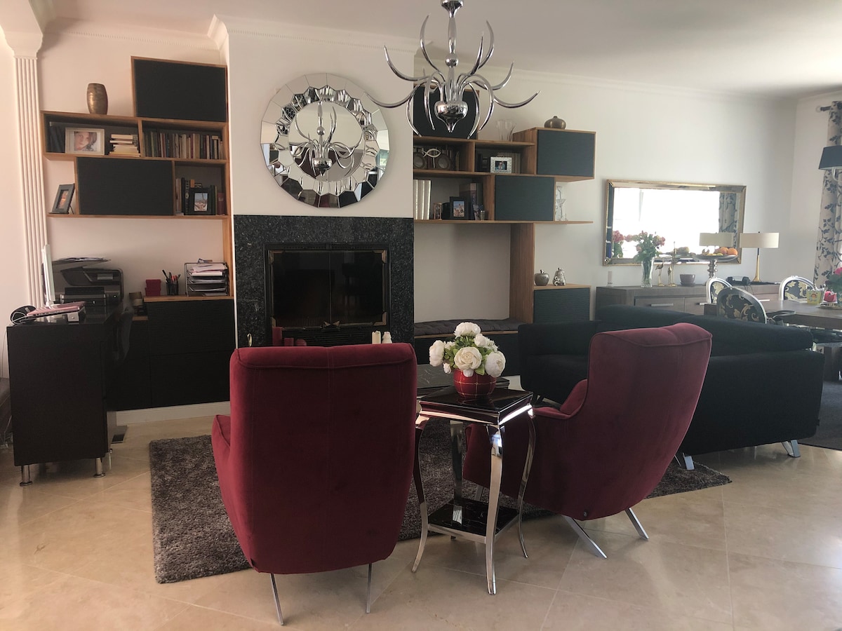A spacious living area features two contemporary burgundy chairs positioned around a glass table. A stylish chandelier hangs above, and a fireplace is set within a black accent wall. Shelving holds various books and decorative items, contributing to the inviting ambiance.