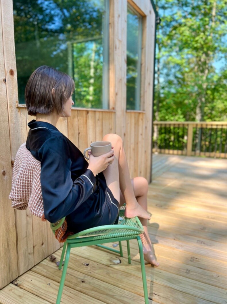 A cozy outdoor seating area features a person sitting on a green chair with legs casually crossed. A mug is held in one hand, while a light blanket drapes over the chair. Sunlight filters through the surrounding trees, creating a serene atmosphere.