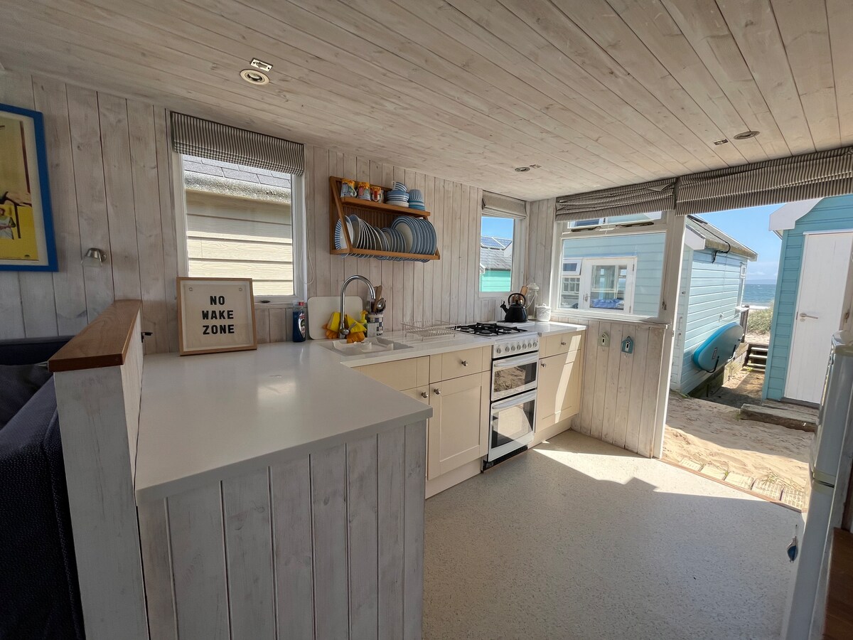 A light-filled kitchenette is equipped with a stove and a fridge, complemented by dishware neatly arranged on shelves. Large windows allow for natural light and offer views of the outdoor area, with the ocean visible in the distance. A comfortable seating area is situated nearby.