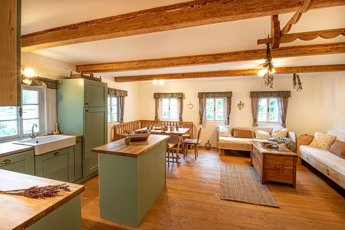 A welcoming living space features rustic wooden beams overhead. A compact kitchen with a farm-style sink is complemented by a central island. A dining area with a wooden table and chairs sits alongside a cozy sitting area with a sofa, creating a communal atmosphere.