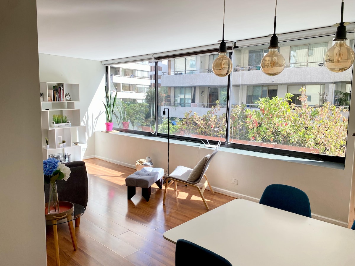 A bright living area features large windows that bring in natural light and offer views of green foliage outside. Minimalist furniture includes a stylish chair and ottoman, alongside a dining table with comfortable chairs. A small bookshelf and decorative plants add to the inviting space.