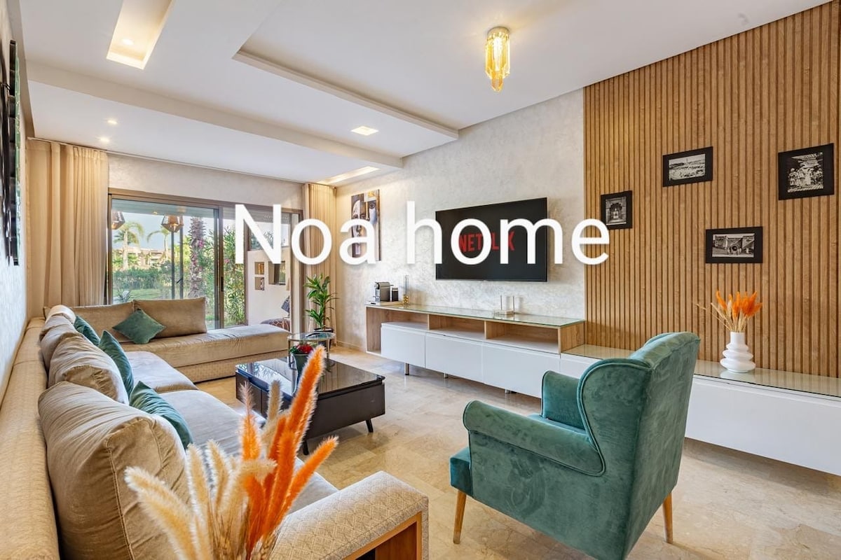 A spacious living area features a comfortable sectional sofa along with a plush accent chair. Natural light enters through large windows, highlighting the elegant decor and wooden accent wall. A flat-screen TV is mounted on the wall, complemented by a modern media console.