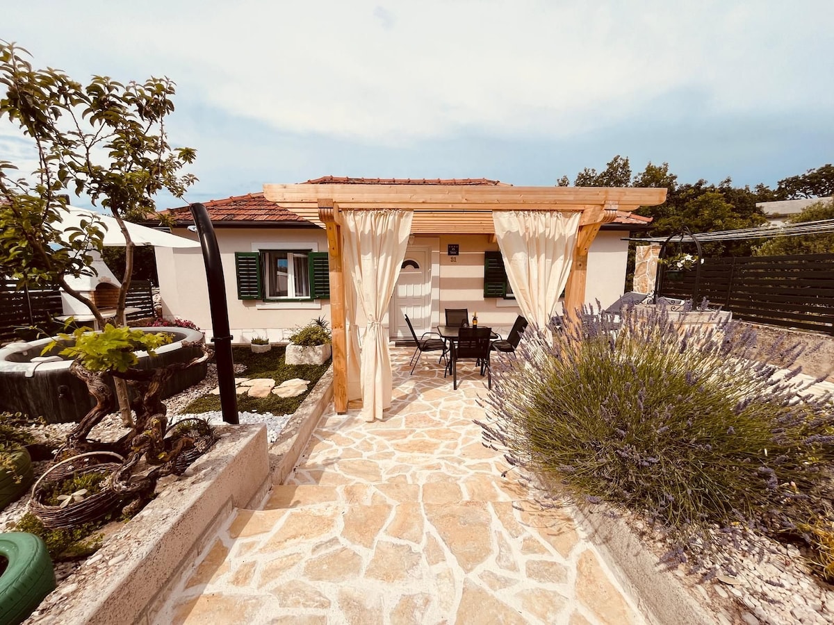 A wooden pergola with flowing curtains frames an outdoor seating area, accompanied by a stone-paved pathway. Lush lavender and ornamental plants enhance the garden's appeal, while a cozy house with green shutters is visible in the background.