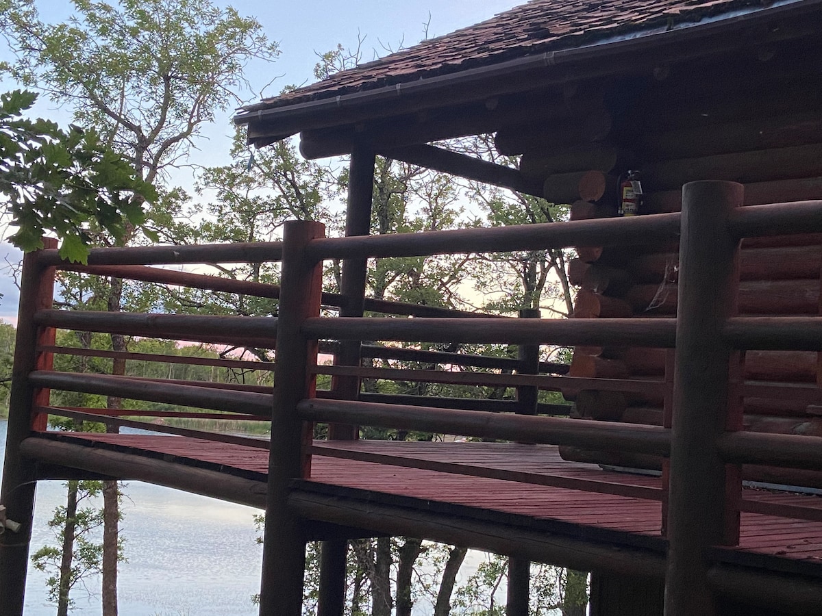A wraparound deck constructed of wood offers views of the surrounding trees and nearby lake. The log cabin structure is partially visible, with railings enhancing the natural setting. Soft light indicates the approach of evening, creating a serene outdoor ambience.