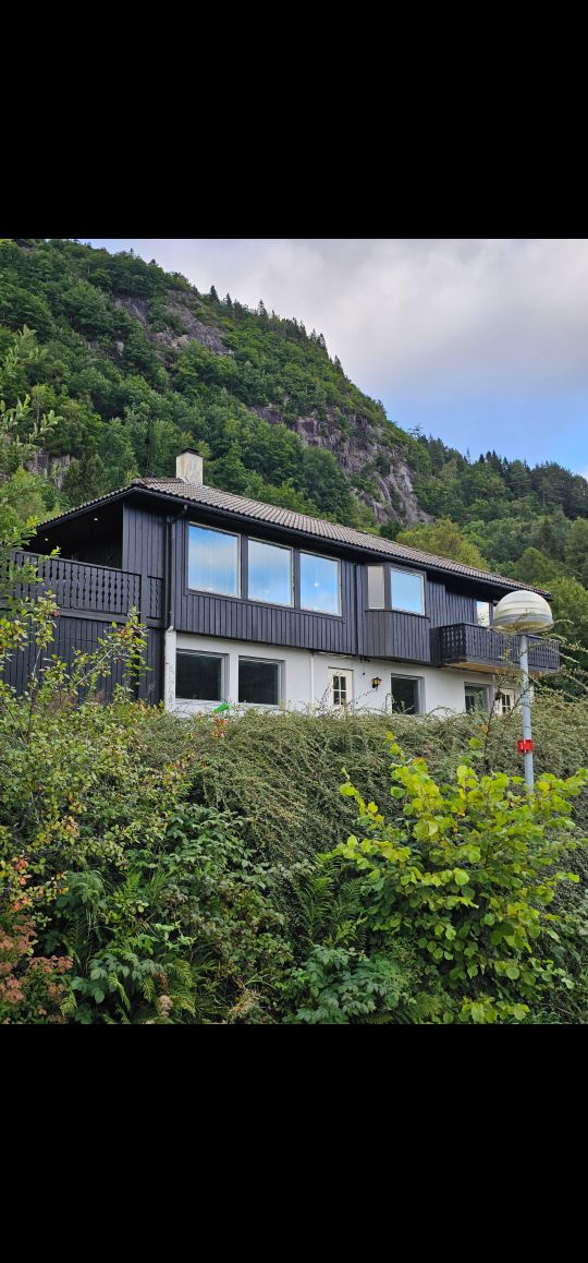 The exterior of a two-story house is surrounded by lush greenery, with a backdrop of mountains. Large windows are visible, allowing natural light to brighten the interior. A balcony extends from the upper level, offering views of the scenic landscape.