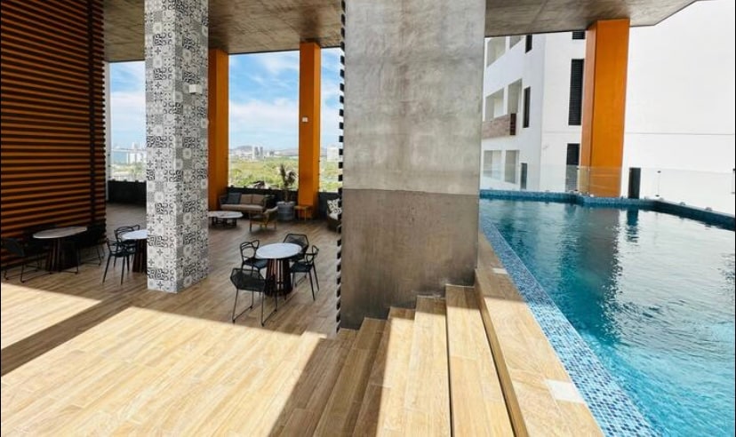An inviting outdoor space features a sleek swimming pool alongside a wooden deck. Several black metal chairs and tables are arranged under a shaded area beside the pool, with vibrant orange and patterned walls providing a modern backdrop.