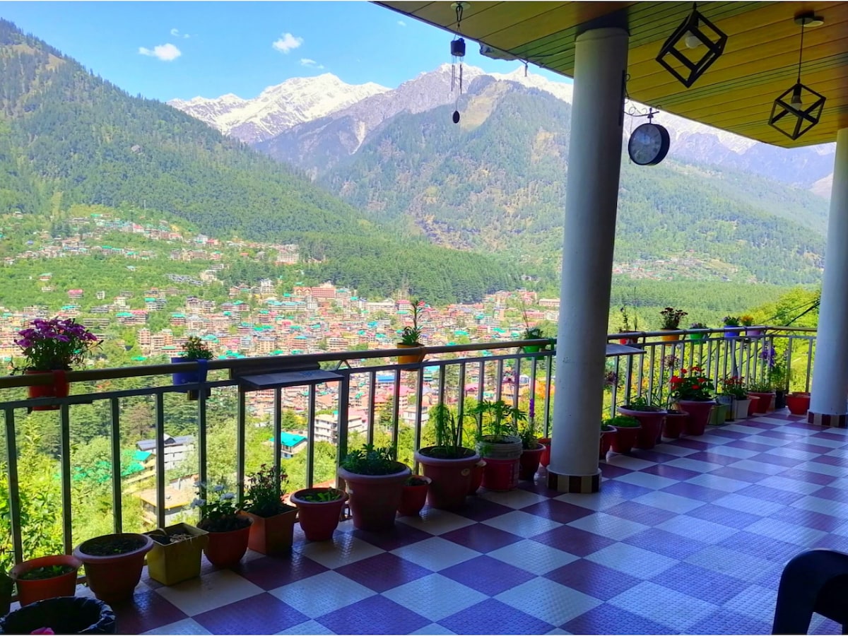 A spacious balcony features colorful pots filled with plants, offering a panoramic view of the mountains in the distance. The valley below is dotted with vibrant rooftops, while natural light enhances the inviting atmosphere of the outdoor space.