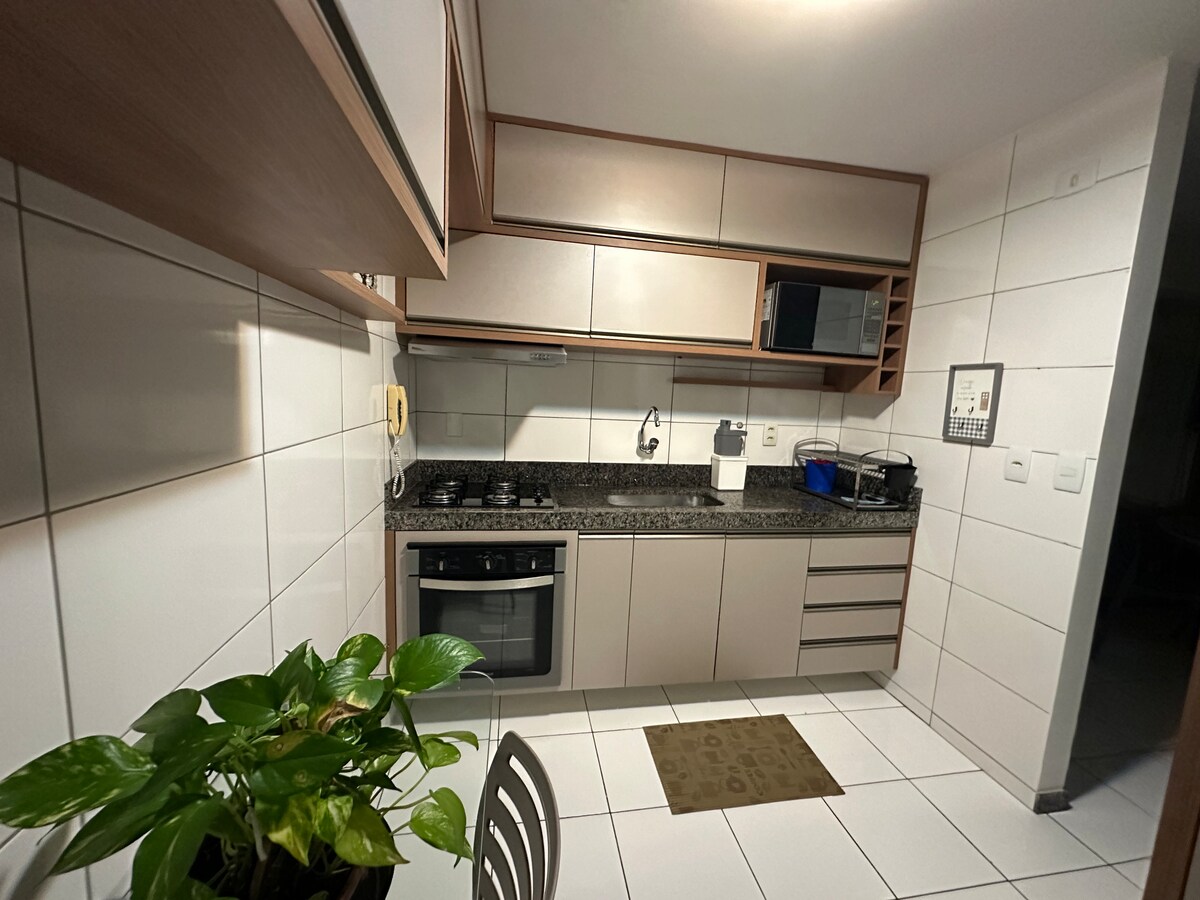 A modern kitchen is presented with sleek cabinetry and granite countertops. Stainless steel appliances, including a stove and microwave, are positioned against tiled walls. A small dining table is visible, accompanied by a potted plant that adds a touch of greenery.