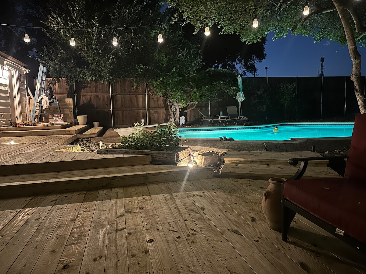 A wooden deck is illuminated by soft string lights, creating a cozy ambiance. A private pool is visible, reflecting evening light. Greenery surrounds the area, with a lounge chair positioned nearby, inviting relaxation. The backdrop is marked by a wooden fence and trees, enhancing the secluded feel.