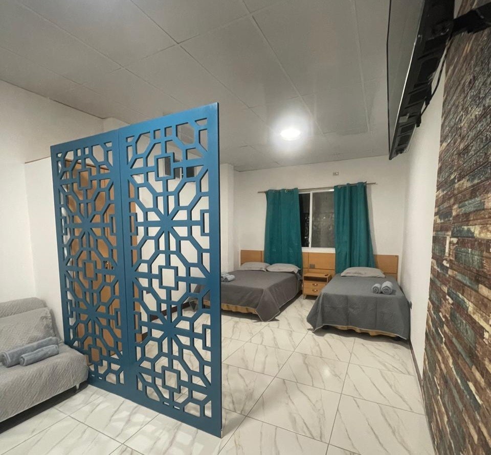 A well-appointed room is divided by a decorative screen, creating a sense of privacy. Two spacious beds are arranged with gray linens and neatly rolled towels. Soft teal curtains frame the window, allowing natural light to enhance the serene environment.