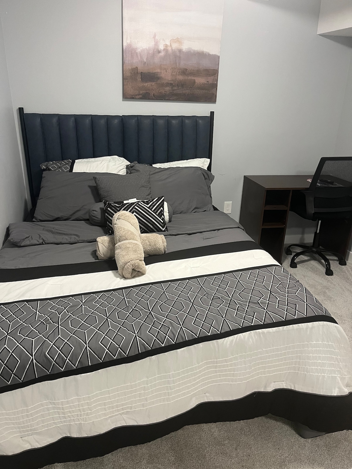 A well-appointed bedroom features a dark upholstered headboard and a neatly arranged bed with grey bedding. A small decorative pillow and rolled towels are placed on the bed. A desk and chair are situated in the corner, against a light blue wall.