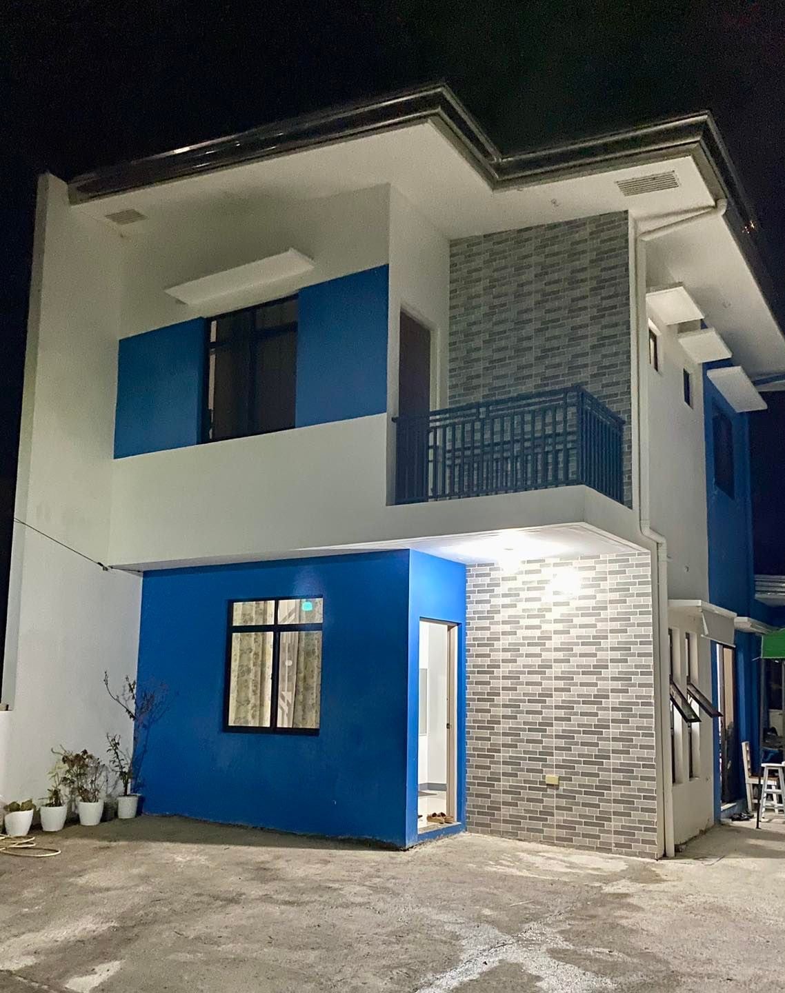The two-story unit features a modern exterior with blue accents and contrasting gray brick. A balcony is visible on the upper floor, complemented by large windows that allow for natural light. The entrance is well-lit, highlighting the surrounding pathway.