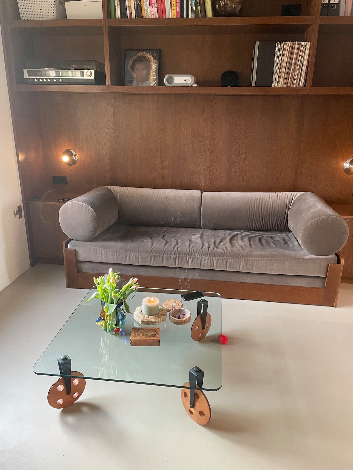 A stylish living area features a cozy, grey couch and a glass coffee table with decorative items. Natural light highlights the space, complemented by wooden shelving filled with books and a projector. Fresh flowers add a touch of greenery to the table.