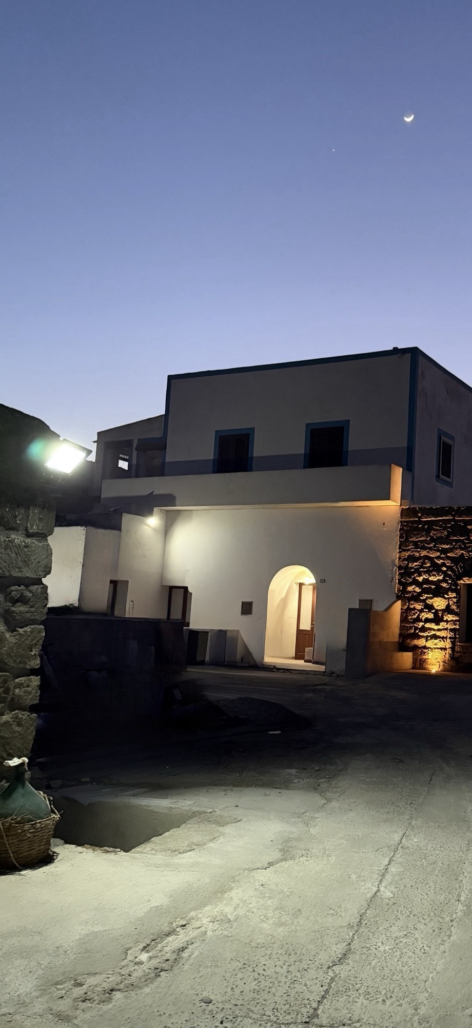 An evening scene captures a tranquil building with soft lighting, creating a welcoming entrance. The structure features a large archway and distinct architectural elements, while the sky transitions from twilight to night, showcasing a hint of the moon above.