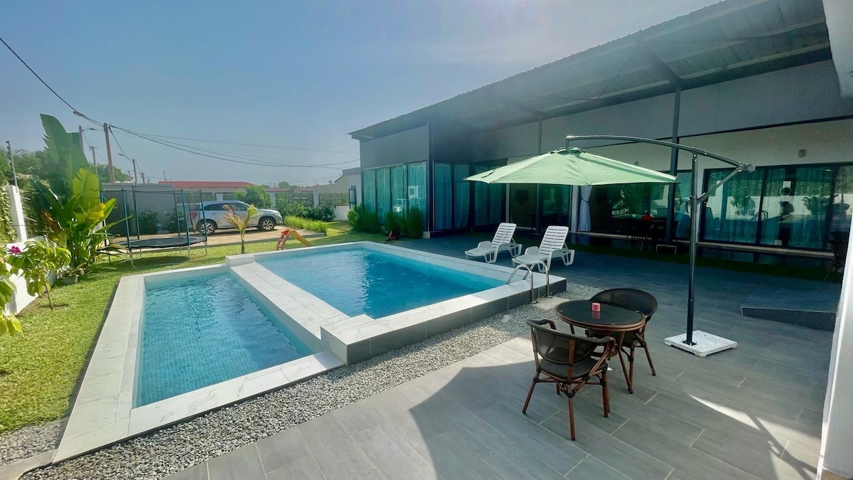 The outdoor area features a swimming pool surrounded by a lawn and paved space. Two lounge chairs are positioned beside the pool. An umbrella shades a small seating arrangement of four chairs and a table, offering a comfortable space for relaxation.