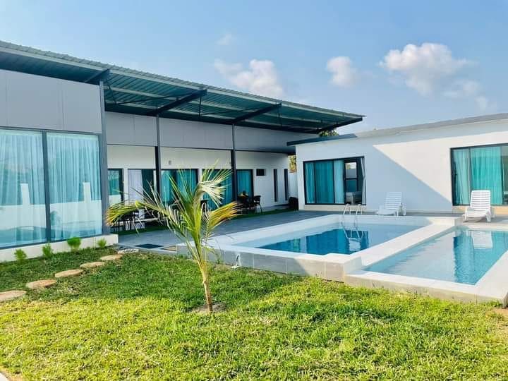 A modern villa is set against a clear blue sky, featuring large glass doors and windows with flowing curtains. A private pool is surrounded by lounge chairs, with well-maintained grass and landscaped paths leading to the entrance.