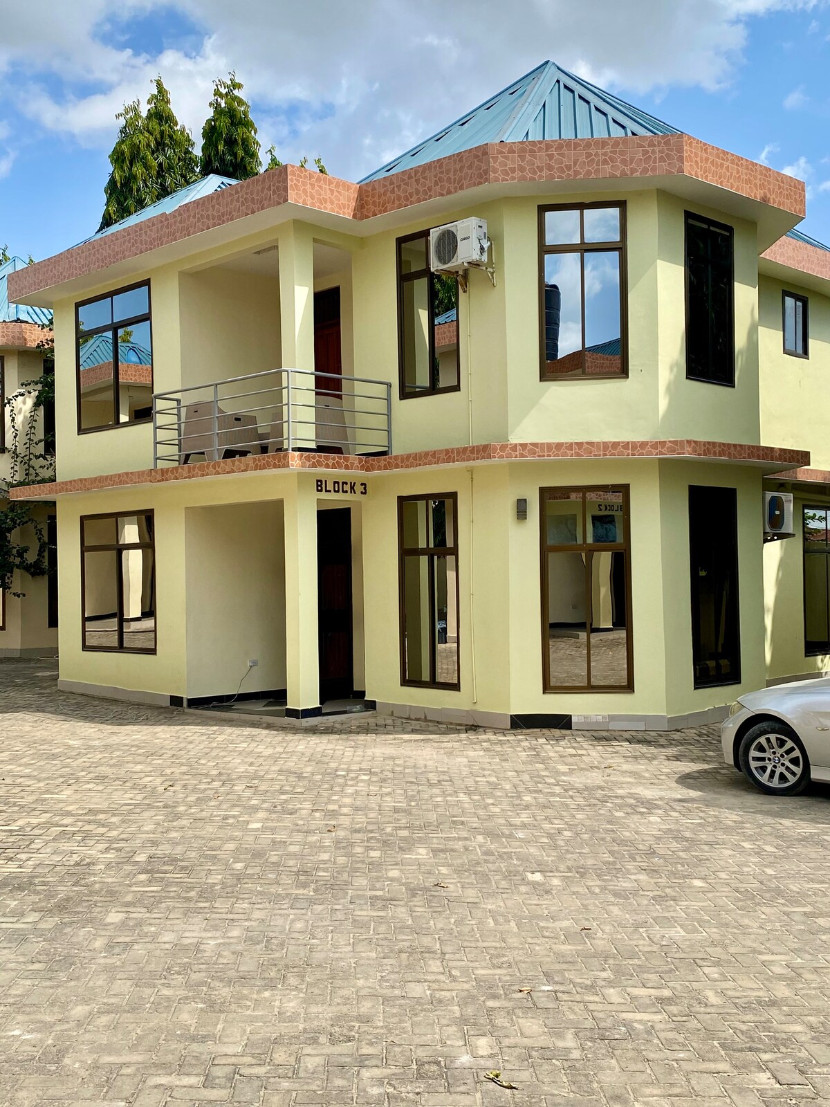The exterior of a two-story home is featured, showcasing large windows that allow natural light to fill the interior. A small balcony is visible on the upper level. The building is painted in a light yellow shade with a blue roof and is situated on a paved area.