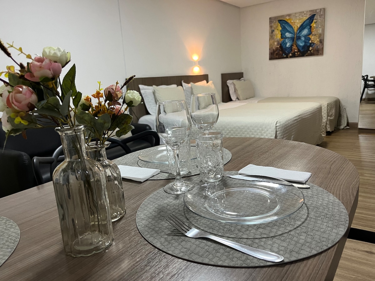 A dining area is presented with a round table set for two, featuring glass plates and cutlery. In the background, a spacious bed with neutral bedding complements a colorful butterfly artwork on the wall. Soft lighting highlights the room's inviting atmosphere.