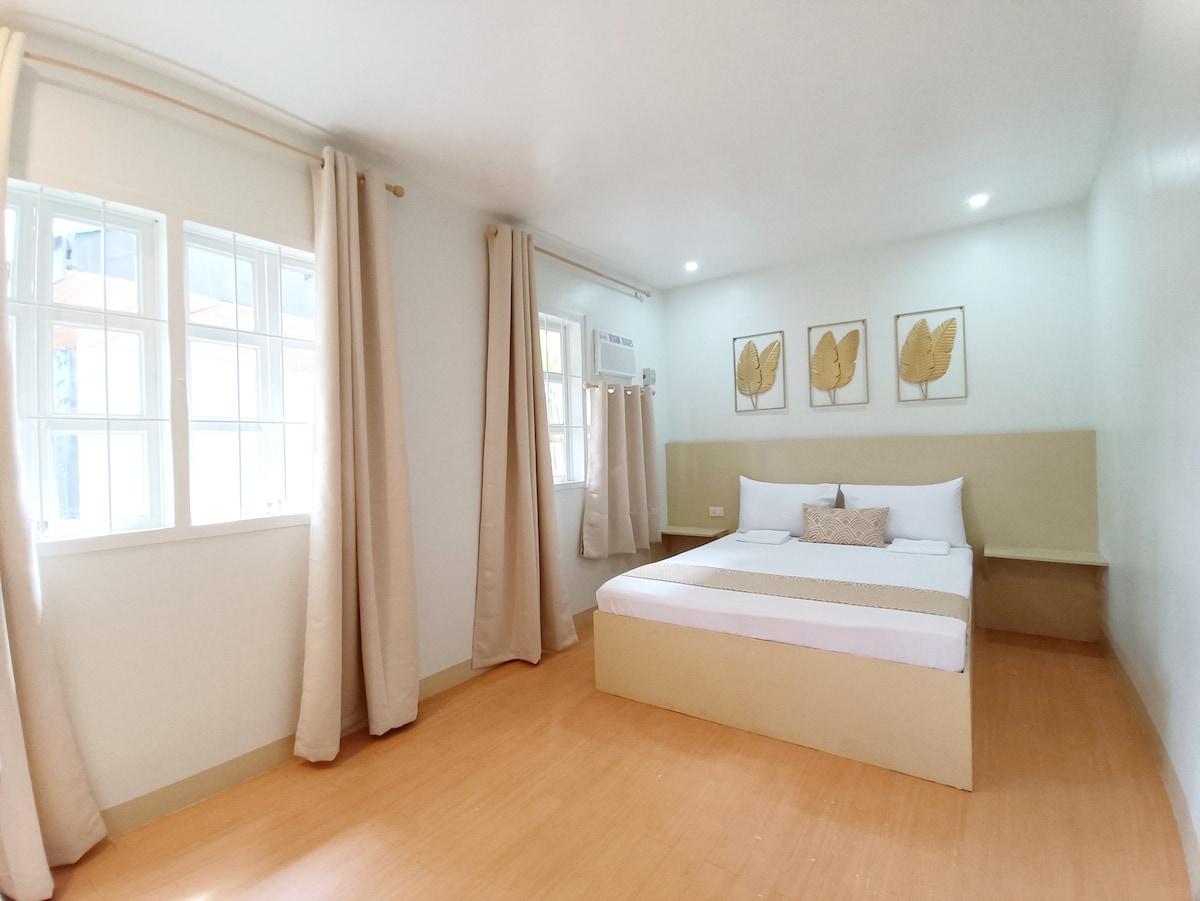 A well-lit guest room features a spacious bed with a neutral-colored headboard. Two windows allow natural light to fill the space, alongside soft curtains. Wall art consisting of leaf motifs complements the serene atmosphere, while a cozy accent pillow adds a touch of comfort.