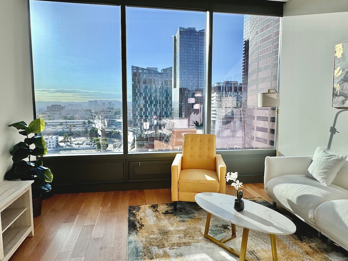 A bright living area features large floor-to-ceiling windows that showcase a view of the downtown skyline. A cozy yellow armchair and a cream-colored sofa are complemented by a neutral area rug and a light wooden coffee table, enhancing the space's inviting feel.